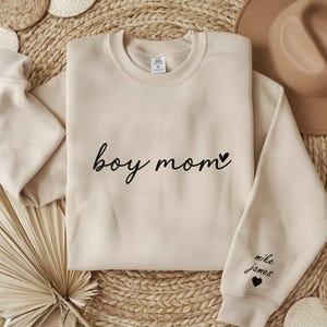 Op de afbeelding: Een beige sweatshirt met de tekst "boy mom" en een klein hartje geprint op de voorkant. De linkermouw heeft de tekst "mike james" en een hartje geprint.