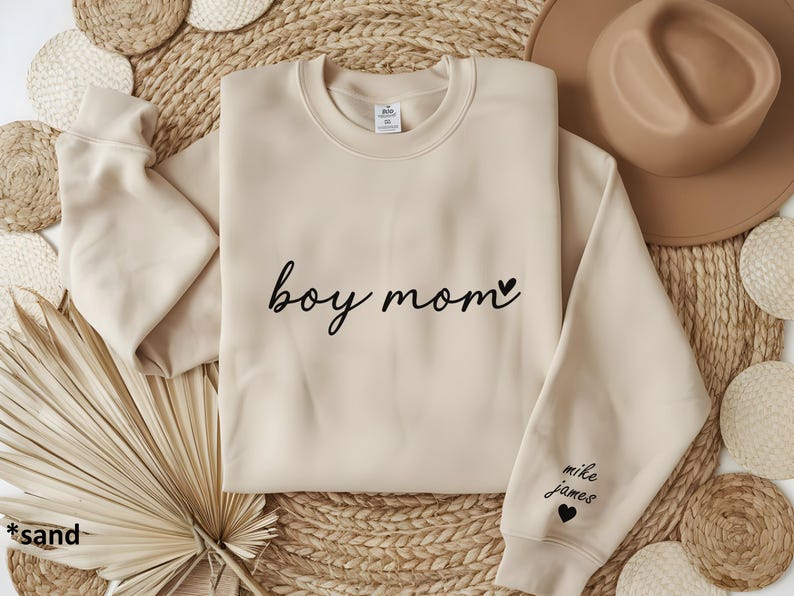 Op de afbeelding: Een beige sweatshirt met de tekst "boy mom" en een klein hartje geprint op de voorkant. De linkermouw heeft de tekst "mike james" en een hartje geprint.