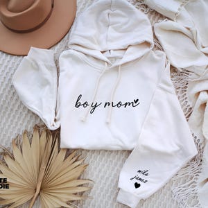 Op de afbeelding: Een witte hoodie met de tekst "boy mom" en een klein hartje op de voorkant. De linkermouw heeft de tekst "mike james" en een klein hartje.
