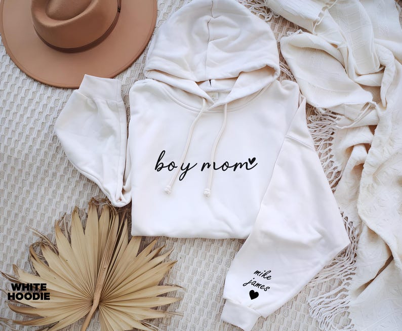 Op de afbeelding: Een witte hoodie met de tekst "boy mom" en een klein hartje op de voorkant. De linkermouw heeft de tekst "mike james" en een klein hartje.