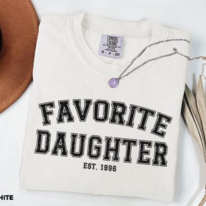 Puede incluir: Una camiseta blanca con la frase "FAVORITE DAUGHTER" en letras negras desgastadas. El texto "EST. 1996" est&aacute; debajo. Un collar plateado con un colgante morado descansa sobre la camiseta doblada.