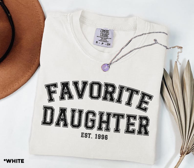 Puede incluir: Una camiseta blanca con la frase "FAVORITE DAUGHTER" en letras negras desgastadas. El texto "EST. 1996" est&aacute; debajo. Un collar plateado con un colgante morado descansa sobre la camiseta doblada.