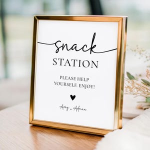 Peut inclure: Un panneau encadré avec un cadre doré et un fond blanc. Le panneau indique "Snack Station" en écriture noire élégante, suivi de "PLEASE HELP YOURSELF. ENJOY!" et d'un symbole de cœur. Les noms "Amy + Adrian" sont en bas.