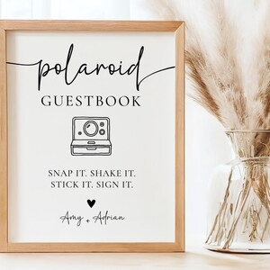 K&ouml;nnte beinhalten: Ein gerahmtes Schild mit den Worten "polaroid GUESTBOOK" in schwarzer Schrift. Darunter befindet sich eine Strichzeichnung einer Polaroid-Kamera. Der Text "SNAP IT. SHAKE IT. STICK IT. SIGN IT." steht unter der Kamera. Die Namen "Amy + Adrian" stehen unten.