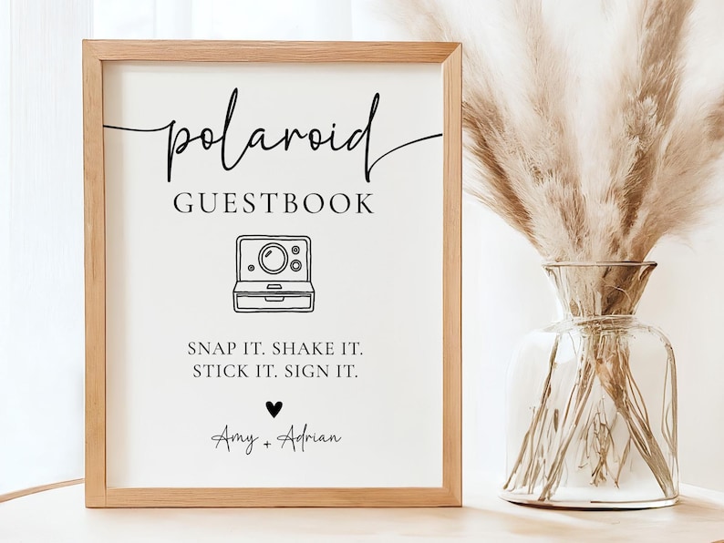 K&ouml;nnte beinhalten: Ein gerahmtes Schild mit den Worten "polaroid GUESTBOOK" in schwarzer Schrift. Darunter befindet sich eine Strichzeichnung einer Polaroid-Kamera. Der Text "SNAP IT. SHAKE IT. STICK IT. SIGN IT." steht unter der Kamera. Die Namen "Amy + Adrian" stehen unten.