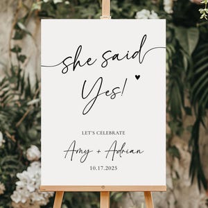 Può includere: Un cartello nuziale color crema su un cavalletto di legno. Il cartello presenta un'elegante scritta nera che recita "she said Yes!" con un simbolo a cuore. Sotto, c'è scritto "LET'S CELEBRATE" e i nomi "Amy + Adrian" con la data "10.17.2025".