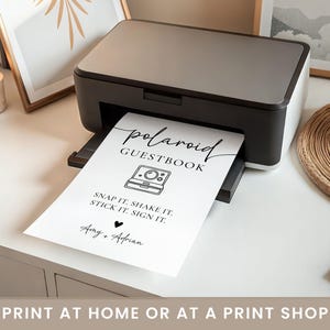 K&ouml;nnte beinhalten: Ein Schwarz-Wei&szlig;-Drucker, der ein "Polaroid G&auml;stebuch"-Schild druckt. Das Schild enth&auml;lt den Text "Snap it. Shake it. Stick it. Sign it." und die Namen "Amy + Adrian". Das Schild hat auch eine Grafik einer Polaroid-Kamera.