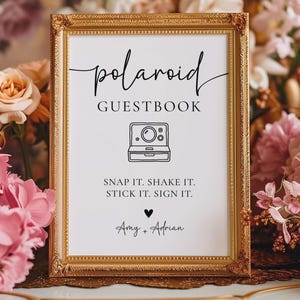 K&ouml;nnte beinhalten: Ein gerahmtes Schild mit goldenem Rand zeigt "polaroid GUESTBOOK" in Schreibschrift. Eine Strichzeichnung einer Vintage-Kamera ist abgebildet. Der Text "SNAP IT. SHAKE IT. STICK IT. SIGN IT." steht darunter, mit den Namen "Amy + Adrian".