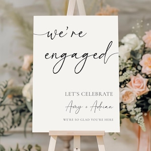 以下が含まれることがあります： 木製のイーゼルに立てられたクリーム色のサイン。黒い筆記体で「we're engaged」と書かれています。その下に「Let's Celebrate」と名前、お祝いのメッセージが書かれています。バラを含むフラワーアレンジメントが見えます。