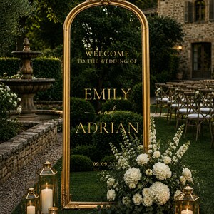 May include: A tall, arched, gold-framed mirror sign featuring the text "WELCOME TO THE WEDDING OF EMILY and ADRIAN 09.09.202X" in gold lettering. The mirror is decorated with a large arrangement of white flowers and two gold lanterns with candles.