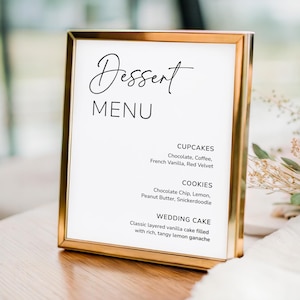 May include: A gold-framed dessert menu with the word "Dessert" in elegant script. The menu lists cupcakes, cookies, and wedding cake flavors, including chocolate, vanilla, and lemon. The sign is on a wooden table.