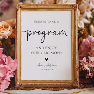 May include: A framed wedding sign with gold trim, displaying the words "PLEASE TAKE A program AND ENJOY OUR CEREMONY." Below, it reads "Ava + Adrian" and a date. The sign is surrounded by pink and peach flowers.