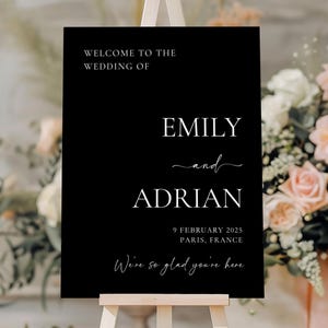 May include: A black wedding welcome sign on a wooden easel. The sign displays the names "EMILY and ADRIAN" with the date "9 FEBRUARY 2025" and location "PARIS, FRANCE". The text "We're so glad you're here" is also included.