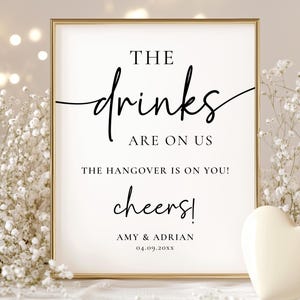 May include: A framed sign with the text "THE drinks ARE ON US" in elegant black script. The sign also includes the text "THE HANGOVER IS ON YOU!" and "cheers!" with names and a date at the bottom. A white heart and flowers are in the background.