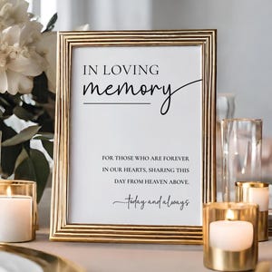 May include: A gold framed sign with the words "IN LOVING memory" in black script. The sign includes the text "FOR THOSE WHO ARE FOREVER IN OUR HEARTS, SHARING THIS DAY FROM HEAVEN ABOVE. Today and always." Gold candle holders and white flowers are in the background.