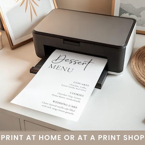 May include: A black and white printer printing a dessert menu. The menu lists cupcakes, cookies, and wedding cake options. The text "Dessert MENU" is at the top of the page in a stylish font. The image also includes a candle, pens, and framed art.