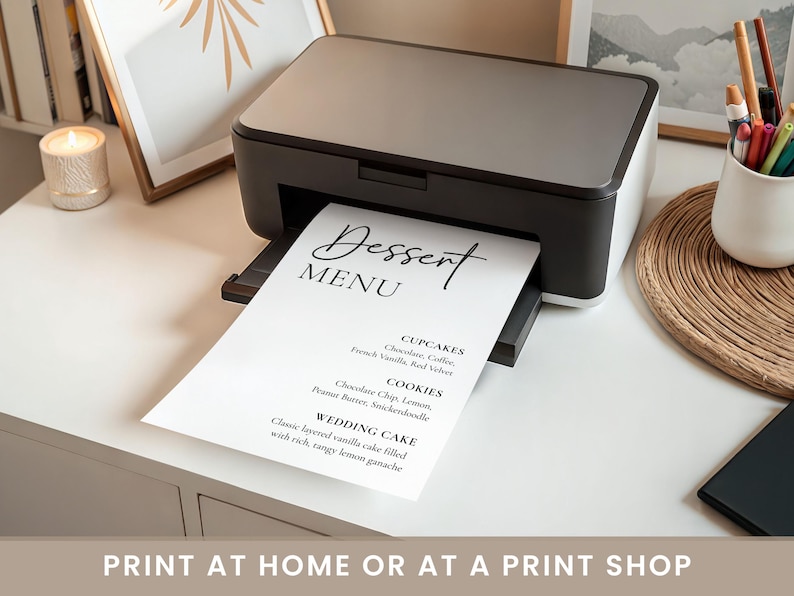 May include: A black and white printer printing a dessert menu. The menu lists cupcakes, cookies, and wedding cake options. The text "Dessert MENU" is at the top of the page in a stylish font. The image also includes a candle, pens, and framed art.