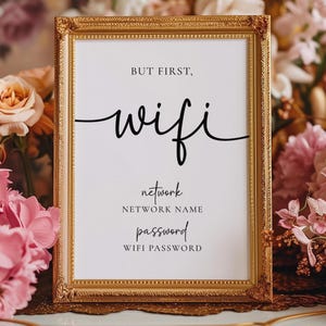 Könnte beinhalten: Ein gerahmtes Schild mit goldfarbenem Rahmen und den Worten "But first, wifi" in schwarzer Schrift. Darunter stehen "network" und "password" mit Platz für den Netzwerknamen und das WLAN-Passwort. Das Schild ist von rosa und pfirsichfarbenen Blumen umgeben.