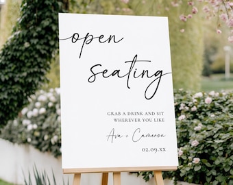 May include: A white sign on a wooden easel with the words "open seating" in elegant black script. Below, it reads "Grab a drink and sit wherever you like" followed by names and a date. The sign is outdoors.