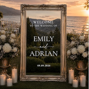 May include: A rectangular mirror with an ornate metallic gold frame featuring white text that reads: WELCOME TO THE WEDDING OF EMILY and ADRIAN 09.09.2026. The mirror reflects a landscape of trees, mountains, and a sunset over the ocean.