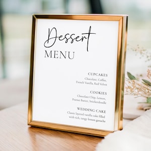 May include: A gold-framed dessert menu with the word "Dessert" in elegant script. The menu lists cupcakes, cookies, and wedding cake flavors, including chocolate, vanilla, and lemon. The sign is on a wooden table.