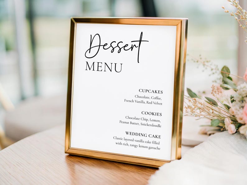 May include: A gold-framed dessert menu with the word "Dessert" in elegant script. The menu lists cupcakes, cookies, and wedding cake flavors, including chocolate, vanilla, and lemon. The sign is on a wooden table.