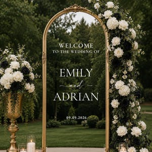 Puede incluir: Un letrero de boda de espejo con marco dorado arqueado y texto blanco que dice Welcome to the wedding of Emily and Adrian 09.09.2026. El espejo está decorado con rosas blancas y negras y follaje, con un jarrón de pedestal dorado a juego.