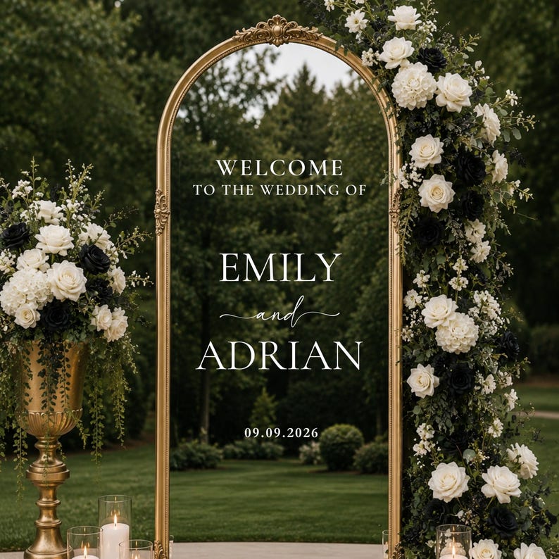 May include: An arched gold-framed mirror wedding sign featuring white text that reads WELCOME TO THE WEDDING OF EMILY and ADRIAN 09.09.2026. The mirror is decorated with white and black roses, greenery, and stands beside a gold pedestal vase.