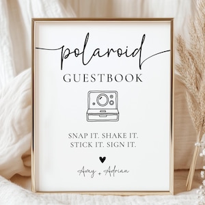 K&ouml;nnte beinhalten: Ein gerahmtes Schild mit den Worten "polaroid GUESTBOOK" in schwarzer Schrift. Darunter eine Strichzeichnung einer Polaroid-Kamera. Der Text "SNAP IT. SHAKE IT. STICK IT. SIGN IT." steht darunter, mit den Namen "Amy + Adrian" am unteren Rand.