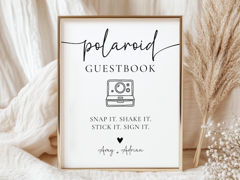 K&ouml;nnte beinhalten: Ein gerahmtes Schild mit den Worten "polaroid GUESTBOOK" in schwarzer Schrift. Darunter eine Strichzeichnung einer Polaroid-Kamera. Der Text "SNAP IT. SHAKE IT. STICK IT. SIGN IT." steht darunter, mit den Namen "Amy + Adrian" am unteren Rand.