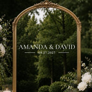 May include: An arched gold metal frame featuring ornate detailing at the top. The center displays the white text AMANDA & DAVID above the date 04.27.2027, flanked by horizontal lines. The frame is decorated with white roses and greenery.