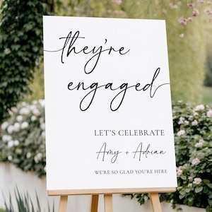 May include: A white sign on a wooden easel with the words "They're engaged" in elegant black script. Below, it reads "Let's Celebrate" and the names "Amy + Adrian". The bottom line says "We're so glad you're here".
