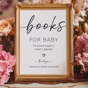 May include: A framed sign with gold trim reads "books FOR BABY" in elegant black script. The sign is surrounded by pink and peach flowers, with text below stating "TO START BABY'S FIRST LIBRARY" and "MARIAH'S BABY SHOWER".