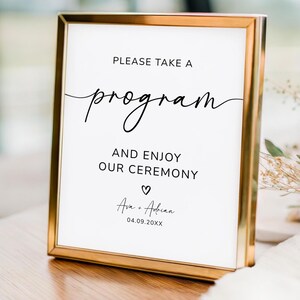 May include: A framed sign with gold trim displays the text "PLEASE TAKE A program AND ENJOY OUR CEREMONY" in black script. Below, the names "Ava + Adrian" and a date are written. The sign is a wedding or event announcement.
