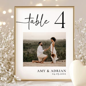 May include: A framed wedding table number with the text "table 4" in a stylish font. The frame contains a photo of a couple, with the names "AMY & ADRIAN" and the date "04.09.20XX" below the photo. The frame is gold.