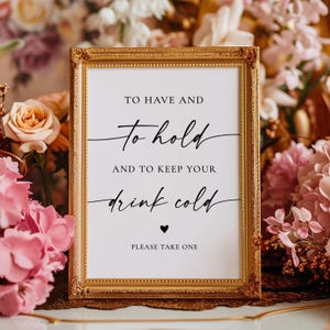 May include: A rectangular sign in a gold ornate frame with white background and black text that reads: TO HAVE AND To hold AND TO KEEP YOUR drink cold ♥ PLEASE TAKE ONE. The sign is surrounded by pink and peach flowers.