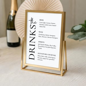 May include: A gold metal framed drinks menu with a white background. The menu lists beer, wine, spirits, and a signature cocktail. The sign is on a white surface. The frame is made of gold metal.