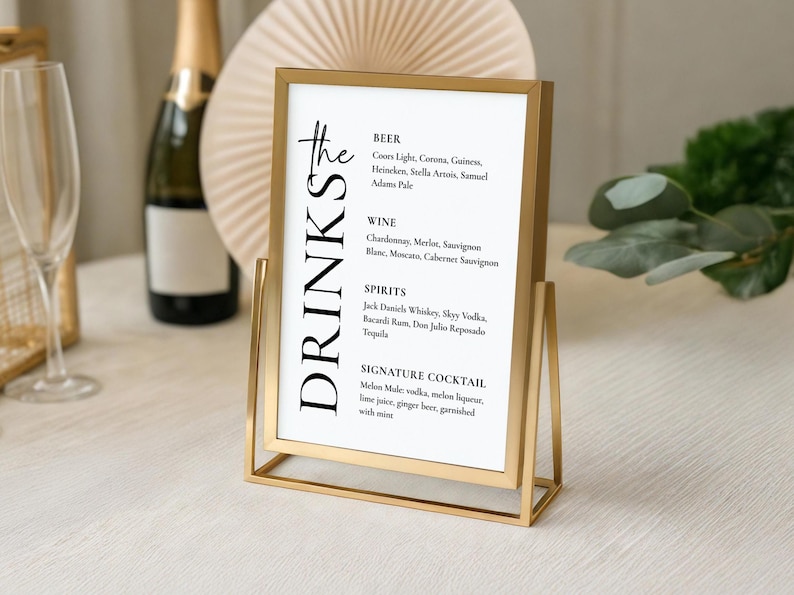 May include: A gold metal framed drinks menu with a white background. The menu lists beer, wine, spirits, and a signature cocktail. The sign is on a white surface. The frame is made of gold metal.