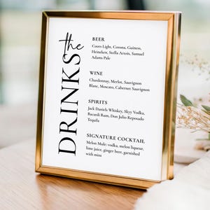 May include: A drinks menu displayed in a gold-framed sign. The sign lists various drinks, including beers, wines, spirits, and a signature cocktail. The word "DRINKS" is written vertically on the left. The sign is placed on a wooden surface.