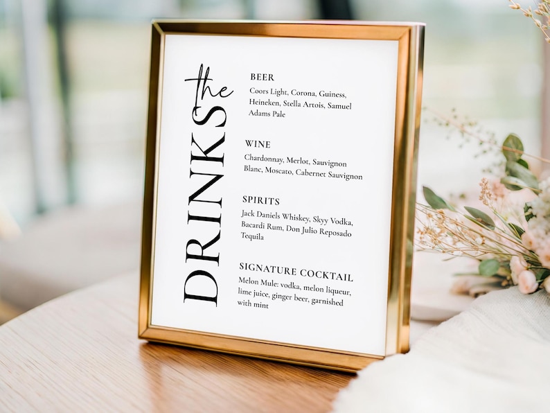 May include: A drinks menu displayed in a gold-framed sign. The sign lists various drinks, including beers, wines, spirits, and a signature cocktail. The word "DRINKS" is written vertically on the left. The sign is placed on a wooden surface.