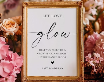 May include: A rectangular gold ornate frame containing a white sign with black text that reads: LET LOVE glow, HELP YOURSELF TO A GLOW STICK AND LIGHT UP THE DANCE FLOOR, a small heart icon, and the names AMY & ADRIAN.