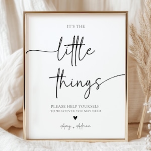 May include: A framed white sign with gold trim features the phrase "It's the little things" in elegant black script. Below, it reads "Please help yourself to whatever you may need." A heart and names complete the design.
