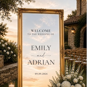 May include: A tall, rectangular mirror with an ornate, metallic gold frame. The glass surface features black printed text that reads: WELCOME TO THE WEDDING OF EMILY and ADRIAN 09.09.2026. The mirror reflects a sunset sky with clouds and light.