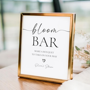 May include: A framed sign with gold trim and white background. The sign reads "bloom BAR" in elegant script, followed by "MAKE A BOUQUET TO TAKE ON YOUR WAY" and "Olivia's Shower". A heart symbol is included.