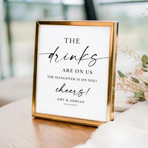 May include: A rectangular gold-toned metal frame containing a white sign with black text that reads: THE drinks ARE ON US THE HANGOVER IS ON YOU! cheers! AMY & ADRIAN 04.29.2027. The sign features a mix of serif and script typography.