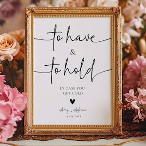 May include: A framed sign with the words "to have & to hold" in elegant black script. Below, it reads "In case you get cold" with names and a date. The frame is ornate gold, surrounded by pink and peach flowers.