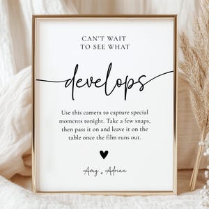 May include: A framed white sign with gold trim features the words "CAN'T WAIT TO SEE WHAT develops" in elegant script. Below, instructions for using a camera are printed. A black heart and names complete the design. The sign is a wedding or event decoration.