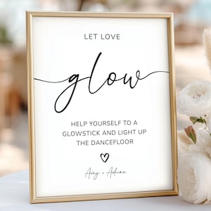 May include: A framed sign with the words "Let Love Glow" in elegant black script. The sign also includes the text "Help yourself to a glowstick and light up the dancefloor" with a heart symbol and names. The frame is gold.