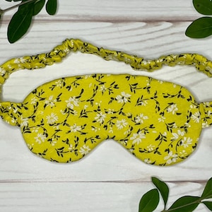 Op de afbeelding: Een gele slaapmasker met een bloemenpatroon van witte bloemen en zwarte stelen. Het masker heeft een bijpassende elastische band. Het slaapmasker is ontworpen om licht te blokkeren voor een betere slaap.