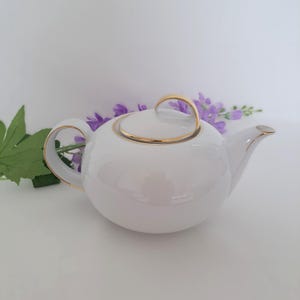 Op de afbeelding: Een witte keramische theepot met een gouden handvat en rand. De theepot heeft een afgeronde vorm en een deksel met een bijpassend gouden handvat. De tuit en de rand van de theepot zijn ook afgewerkt met goud. De achtergrond bevat paarse bloemen en groene bladeren.